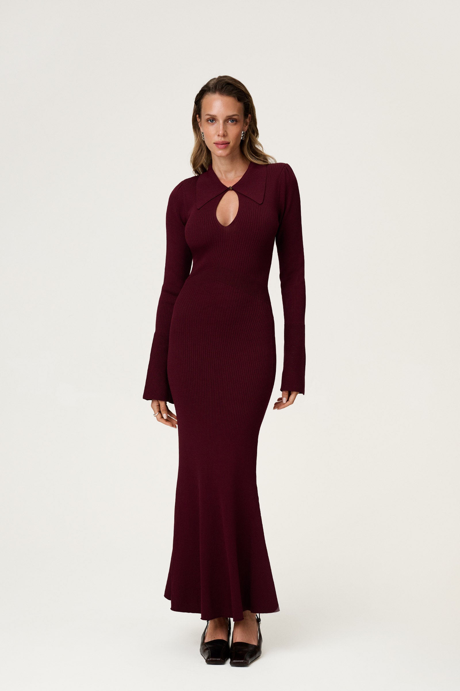 Woman wearing the Basel dress in burgundy, a knitted maxi with a collared cut-out neckline, decorative seams, and a flared hem. Finished with long sleeves and wide cuffs. image