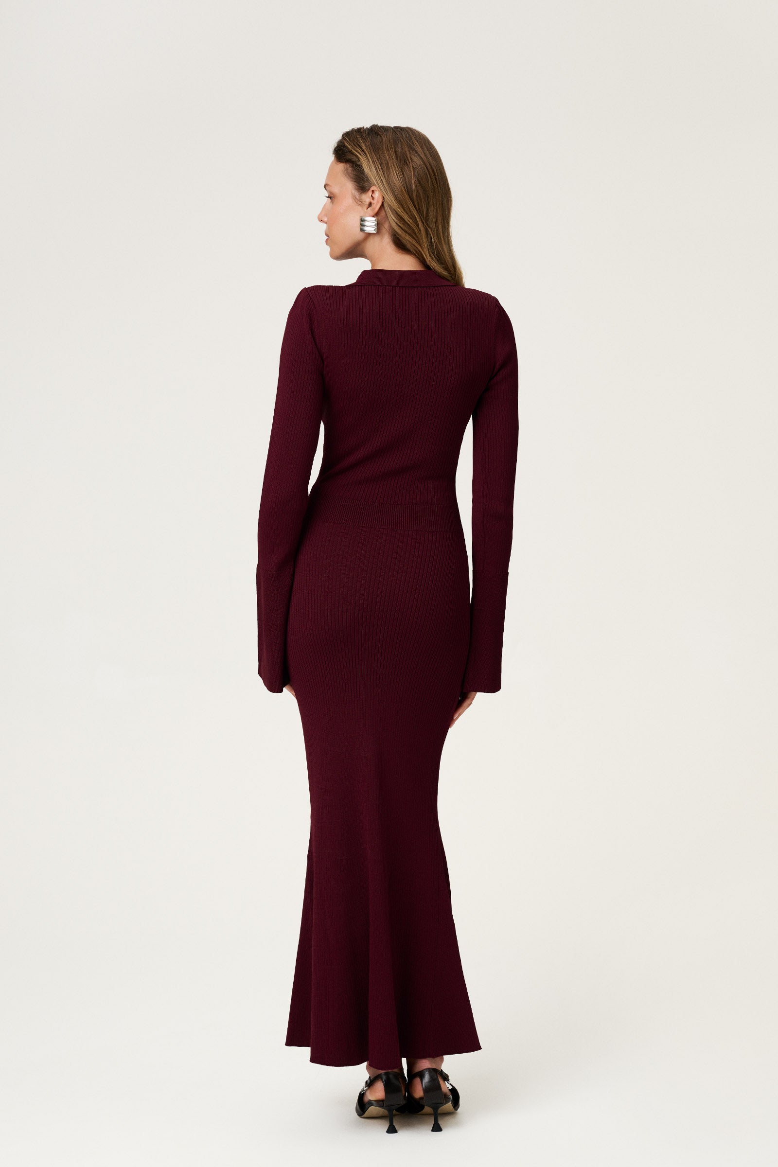 Woman wearing the Basel dress in burgundy, a knitted maxi with a collared cut-out neckline, decorative seams, and a flared hem. Finished with long sleeves and wide cuffs. image