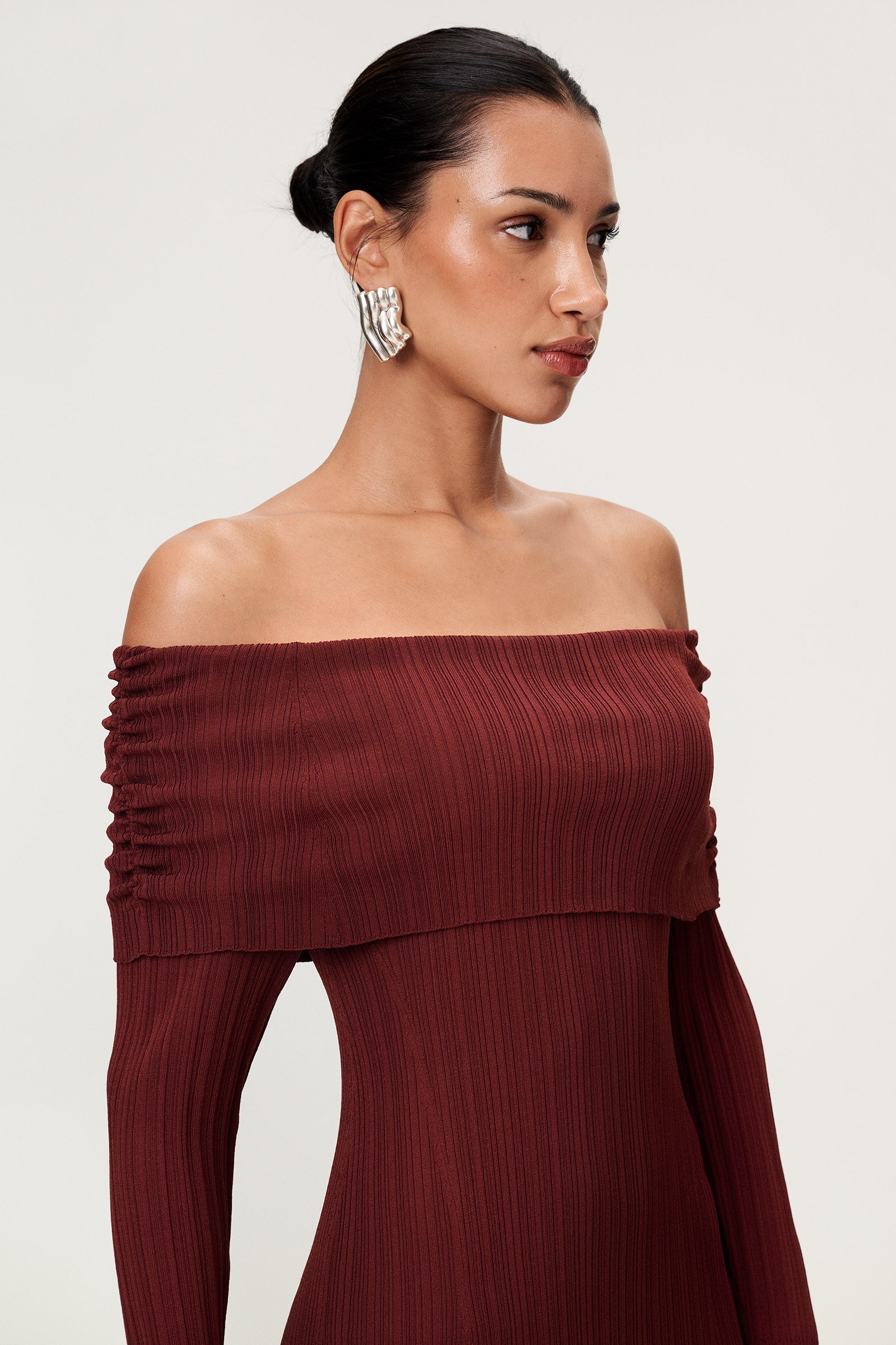 Woman wearing the Izano dress in burgundy, a knitted off-shoulder maxi dress with ruched panel detailing, soft draping, and a flowing, cozy silhouette. image
