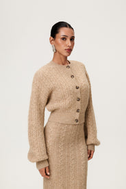 Woman wearing the Merano cardigan in beige melange, a cropped knit with puff sleeves, dropped shoulders, fitted cuffs, and a ribbed waist with button closure. thumbnail image