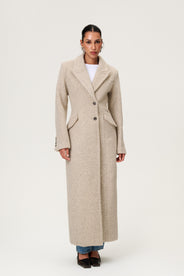 Woman wearing the Stockholm coat in beige melange. A full-length, hourglass coat with a cinched waist, removable suede belt, and structured shoulders. Finished with front pockets and a fully lined interior for warmth and comfort. thumbnail image
